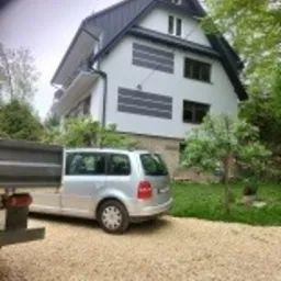 Dwukondygnacyjny dom z jasną elewacją, częściowo ocieplony szarymi panelami, widoczny z poziomu żwirowego podjazdu, obok zaparkowany srebrny samochód dostawczy.