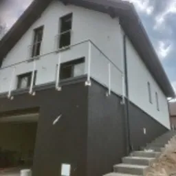 Dwukondygnacyjny dom z częściowo otynkowaną elewacją w kolorze białym, z widocznym czarnym podpiwniczeniem i schodami prowadzącymi do wejścia, teren wokół w trakcie prac wykończeniowych.