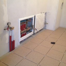 Przemysław Świderski - Układanie korkowych płytek podłogowych w łazience, widoczny rozdzielacz ogrzewania podłogowego w zabudowie, rury doprowadzające wodę z izolacją i manometr.