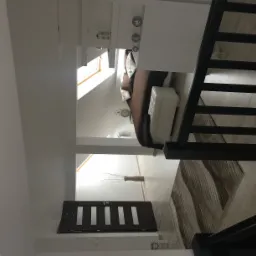 Jasne poddasze z widokiem na łóżko, schody z czarną balustradą i drzwi do toalety z minimalistycznym wzorem.