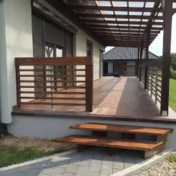 Zadaszony taras z drewnianą konstrukcją, brązowymi płytkami podłogowymi i drewnianą balustradą. Widoczne dwustopniowe drewniane schody i szara kostka brukowa z białym obrzeżem.