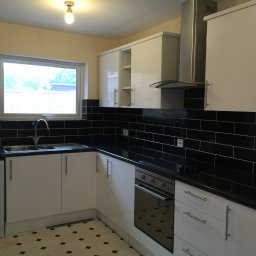 Przemysław Świderski - Nowoczesna kuchnia z białymi szafkami, czarnym blatem i płytkami, z pralką wbudowaną pod blatem, oświetlona pojedynczą żarówką.