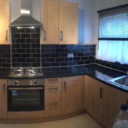 Przemysław Świderski - Jasna kuchnia z drewnianymi szafkami, czarnym blatem i czarnymi płytkami nad blatem, okapem nad kuchenką gazową i zlewozmywakiem pod oknem z żaluzjami i firanką.