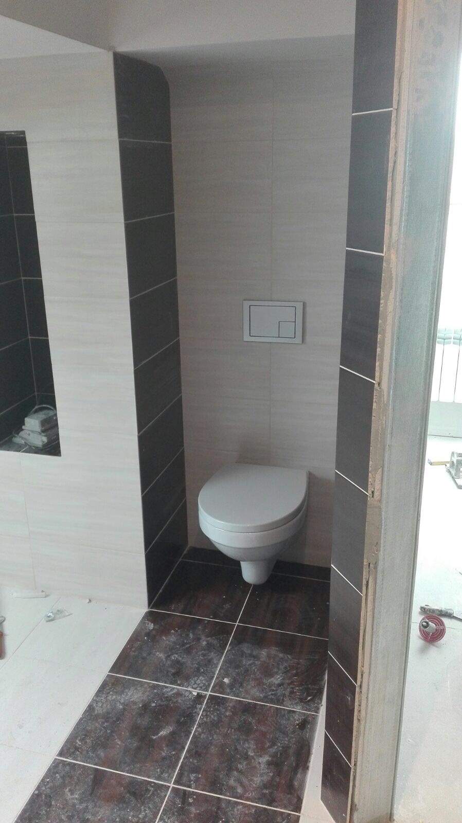 Zamontowana toaleta wisząca z przyciskiem spłukującym na ścianie, obłożona płytkami w odcieniach szarości i brązu, widoczne niedociągnięcia w wykończeniu przy framudze drzwi.