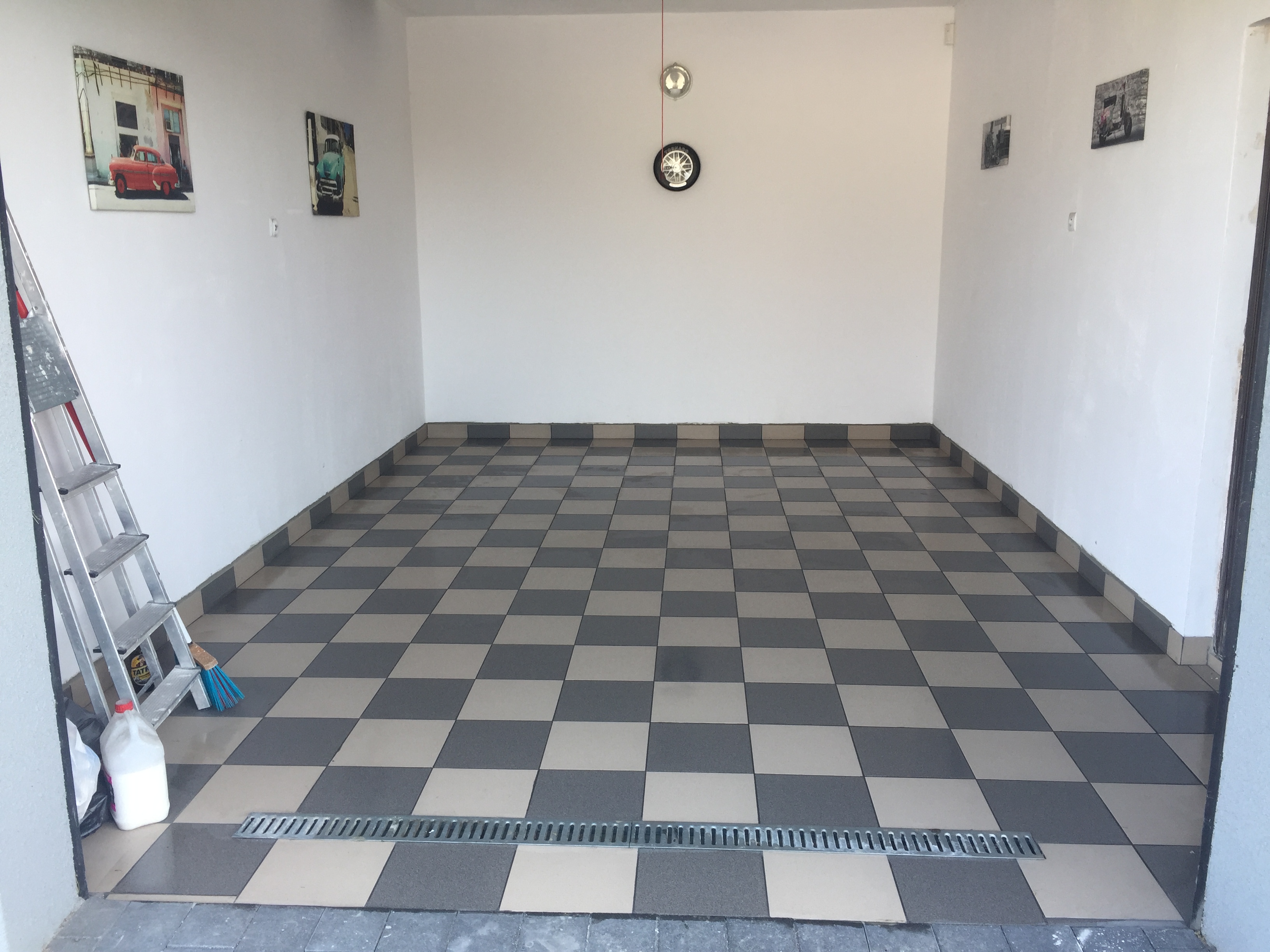 Szachownica z szarych i beżowych płytek na podłodze garażu, z odpływem liniowym, drabina aluminiowa i pojemnik z płynem czyszczącym w rogu, ściany białe z obrazkami samochodów i zegarem.