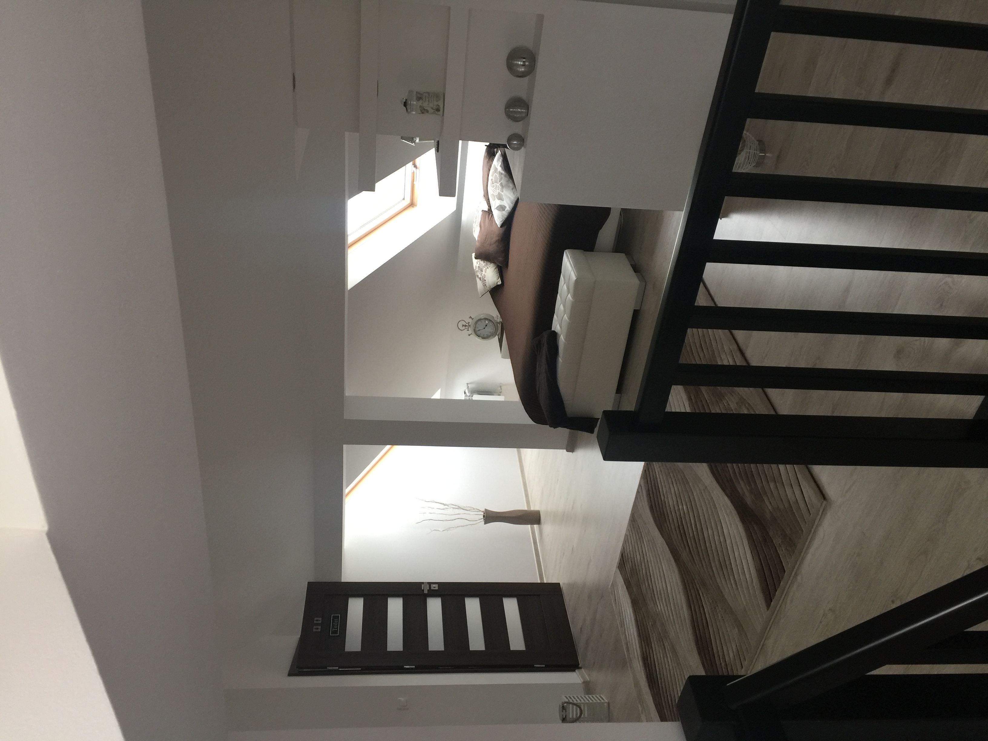 Jasne poddasze z widokiem na łóżko, schody z czarną balustradą i drzwi do toalety z minimalistycznym wzorem.