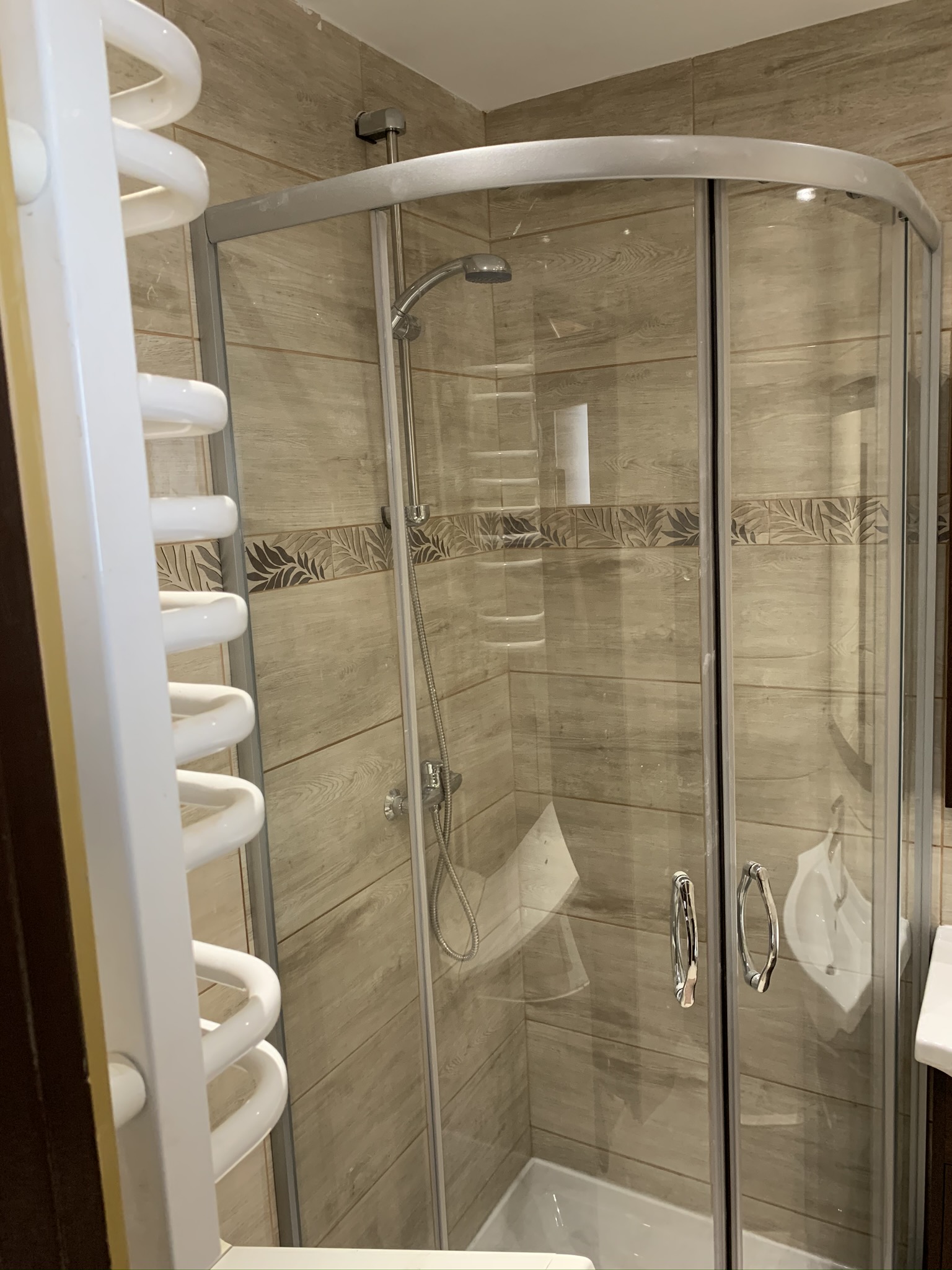 Kabina prysznicowa z chromowanymi uchwytami i deszczownicą, obok biały grzejnik drabinkowy na tle beżowych płytek ceramicznych z dekoracyjnym pasem z motywem liści.