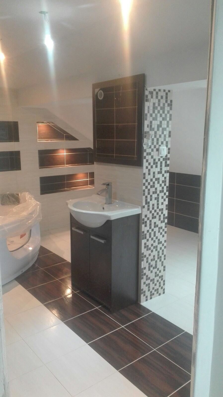 Wykończona łazienka z umywalką narożną na szafce w kolorze ciemnego drewna, płytki podłogowe biało-brązowe, ściany z wnękami wyłożonymi brązowymi płytkami, kolumna z mozaiki czarno-białej, toaleta...