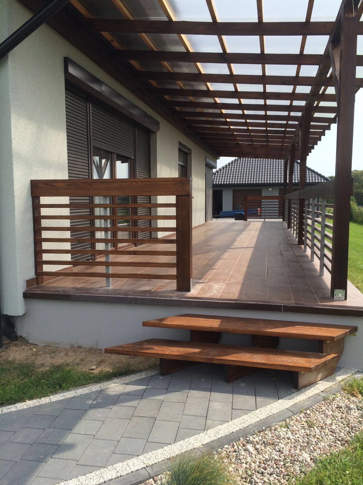 Zadaszony taras z drewnianą konstrukcją, brązowymi płytkami podłogowymi i drewnianą balustradą. Widoczne dwustopniowe drewniane schody i szara kostka brukowa z białym obrzeżem.