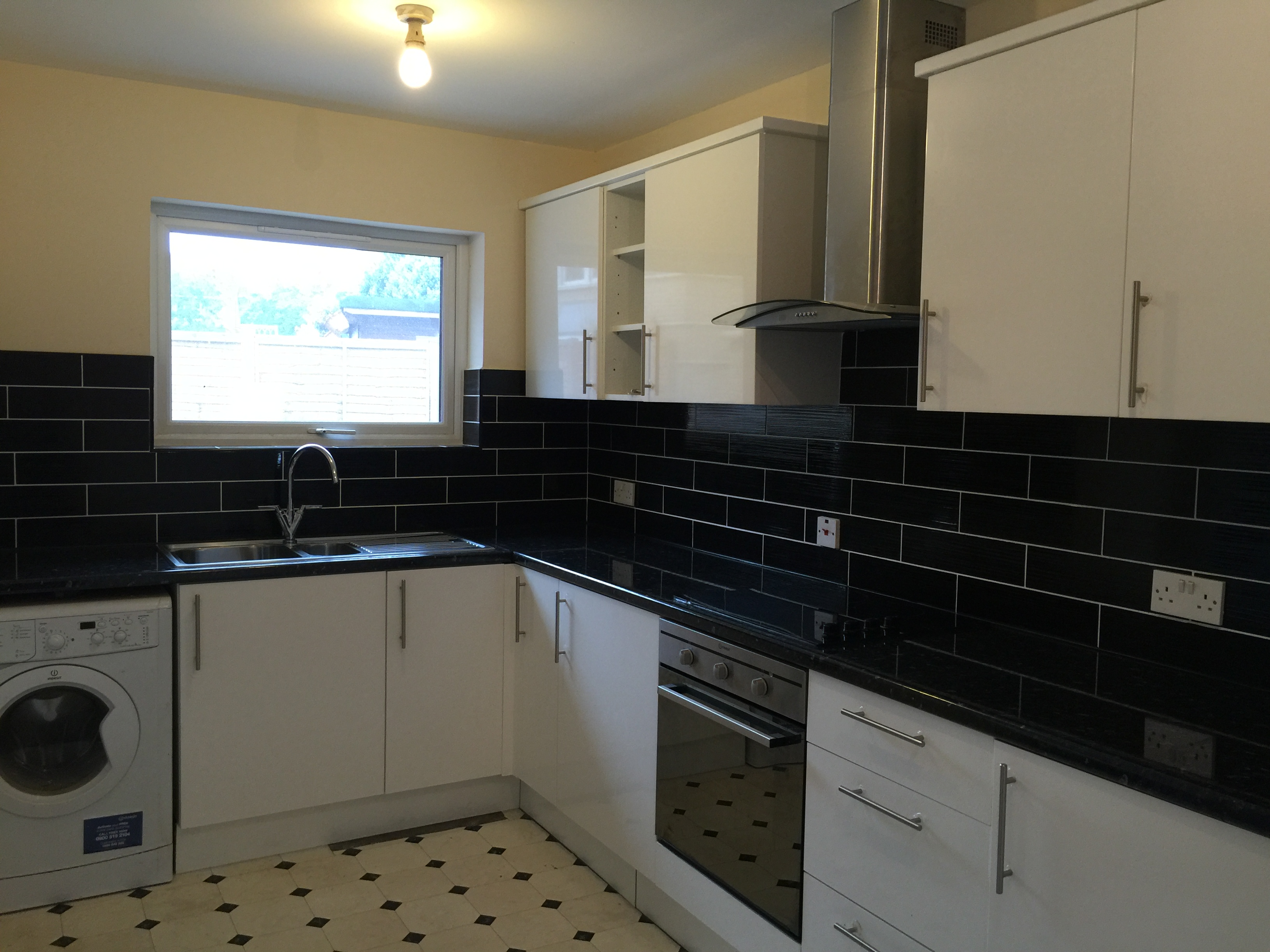 Nowoczesna kuchnia z białymi szafkami, czarnym blatem i płytkami, z pralką wbudowaną pod blatem, oświetlona pojedynczą żarówką.