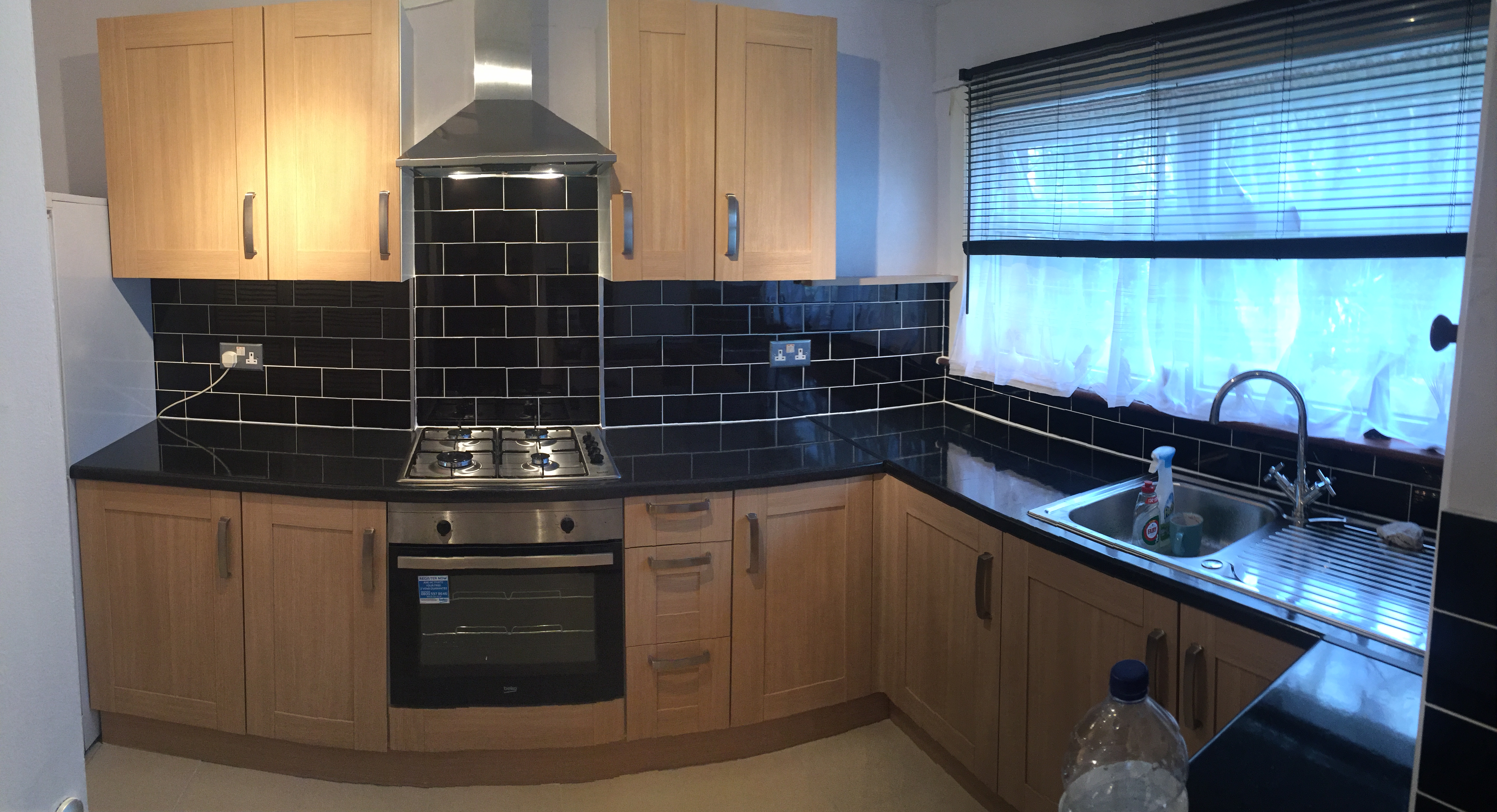 Jasna kuchnia z drewnianymi szafkami, czarnym blatem i czarnymi płytkami nad blatem, okapem nad kuchenką gazową i zlewozmywakiem pod oknem z żaluzjami i firanką.