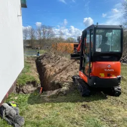 Pomarańczowa mini koparka Kubota obok wykopu w ziemi przy białej ścianie budynku, w tle drzewa i błękitne niebo z chmurami, na trawie leżą robocze ubrania.