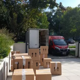 Ciężarówka transportowa z otwartymi drzwiami i stosem kartonowych pudeł oznaczonych strzałkami na chodniku przed budynkiem, w tle drzewa i błękitne niebo.