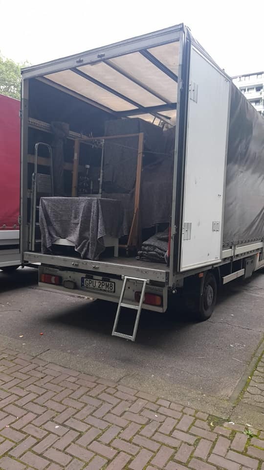 Otwarta paka ciężarówki załadowana meblami przykrytymi szarą tkaniną, drabinka opuszczona na chodnik z kostki brukowej, widoczna tablica rejestracyjna GPU 2PM8.