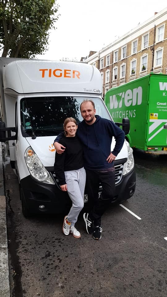 Uśmiechnięta para stoi przed białym busem firmy TIGER Twin Cab z pomarańczowym napisem na tle ulicy z budynkami mieszkalnymi i zieloną furgonetką WREN KITCHENS w tle.