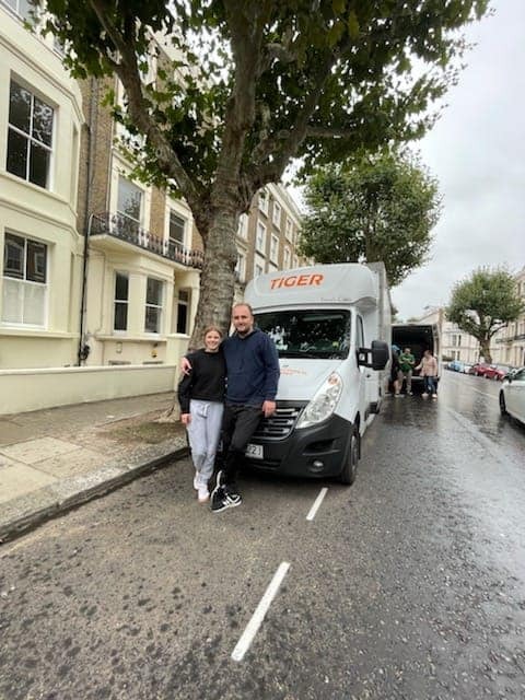 Uśmiechnięta para stoi przed białą furgonetką z napisem 'TIGER' na tle miejskiej ulicy w deszczowy dzień, z otwartymi drzwiami furgonetki i osobami w tle.