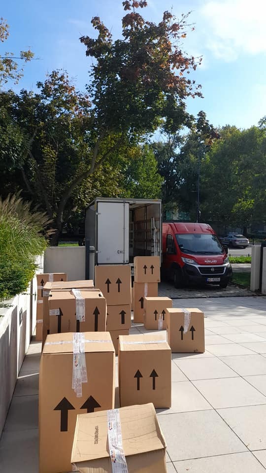 Ciężarówka transportowa z otwartymi drzwiami i stosem kartonowych pudeł oznaczonych strzałkami na chodniku przed budynkiem, w tle drzewa i błękitne niebo.