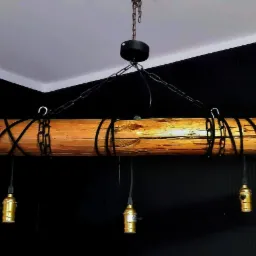 Drewniana belka jako lampa wisząca z trzema złotymi oprawkami żarówek, zawieszona na czarnych łańcuchach w pomieszczeniu z czarną ścianą.