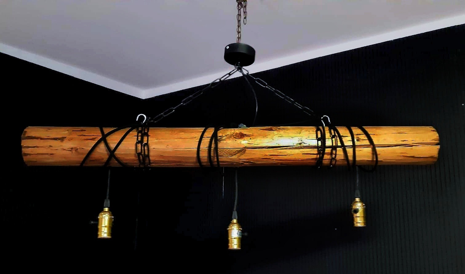 Drewniana belka jako lampa wisząca z trzema złotymi oprawkami żarówek, zawieszona na czarnych łańcuchach w pomieszczeniu z czarną ścianą.
