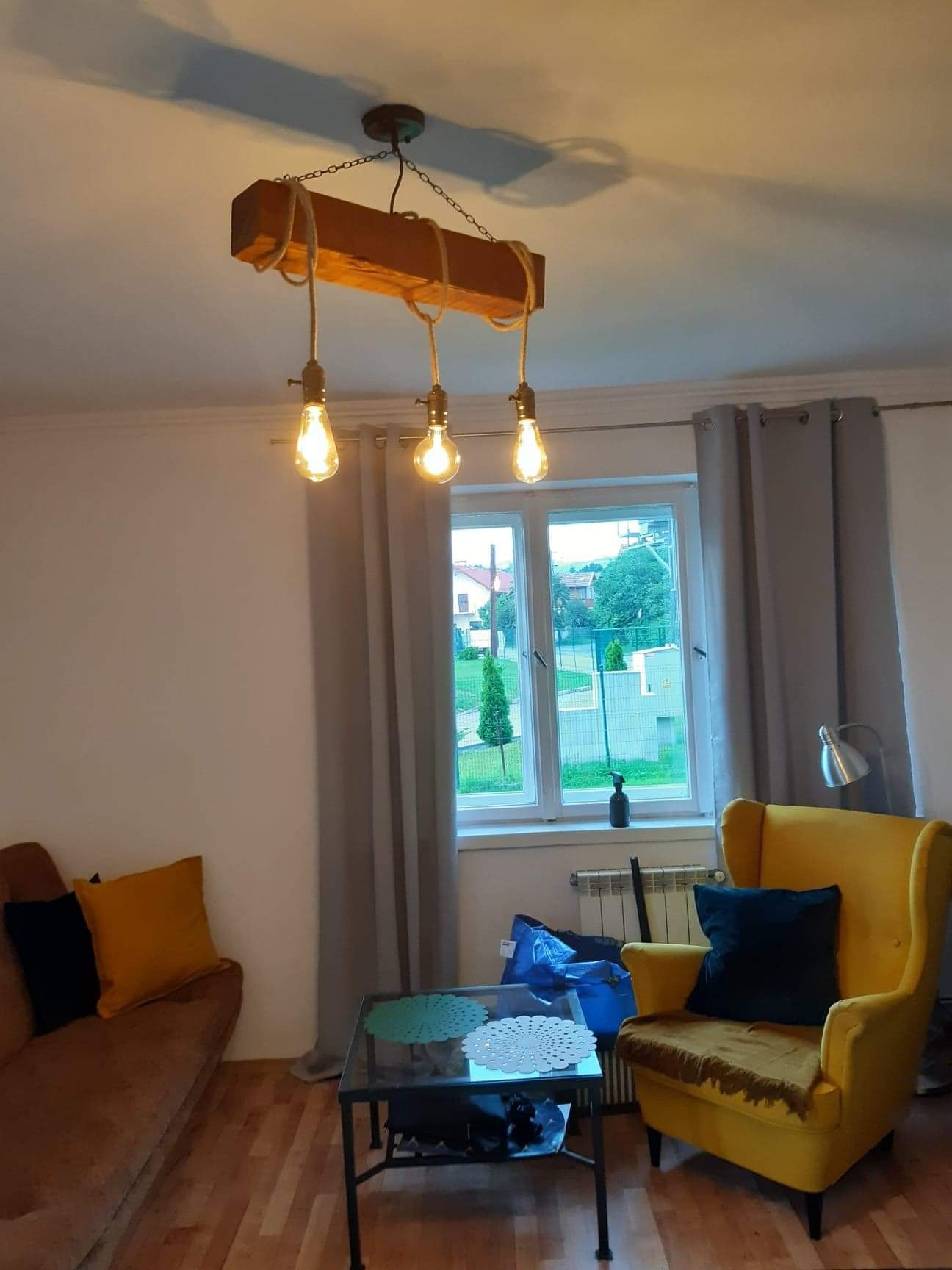 Rustykalna lampa wisząca z drewnianej belki i sznurów z trzema żarówkami Edisona w salonie z żółtym fotelem i brązową kanapą.