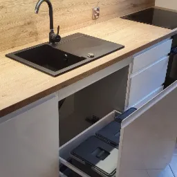 Nowoczesna kuchnia z drewnianym blatem, czarnym zlewem i wysuwanym koszem na śmieci w szafce.