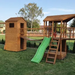 Drewniany plac zabaw z wieżą, zjeżdżalnią i huśtawką na zielonym trawniku, w tle domy i drzewa.