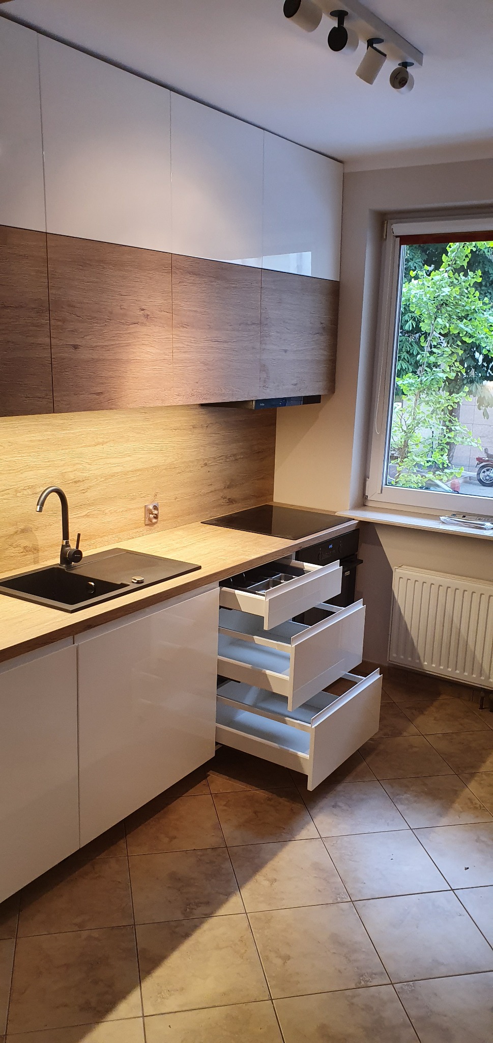 Nowoczesna kuchnia z białymi szafkami i drewnianymi akcentami, czarnym zlewem i trzema wysuniętymi szufladami, widok na okno z zielenią na zewnątrz.