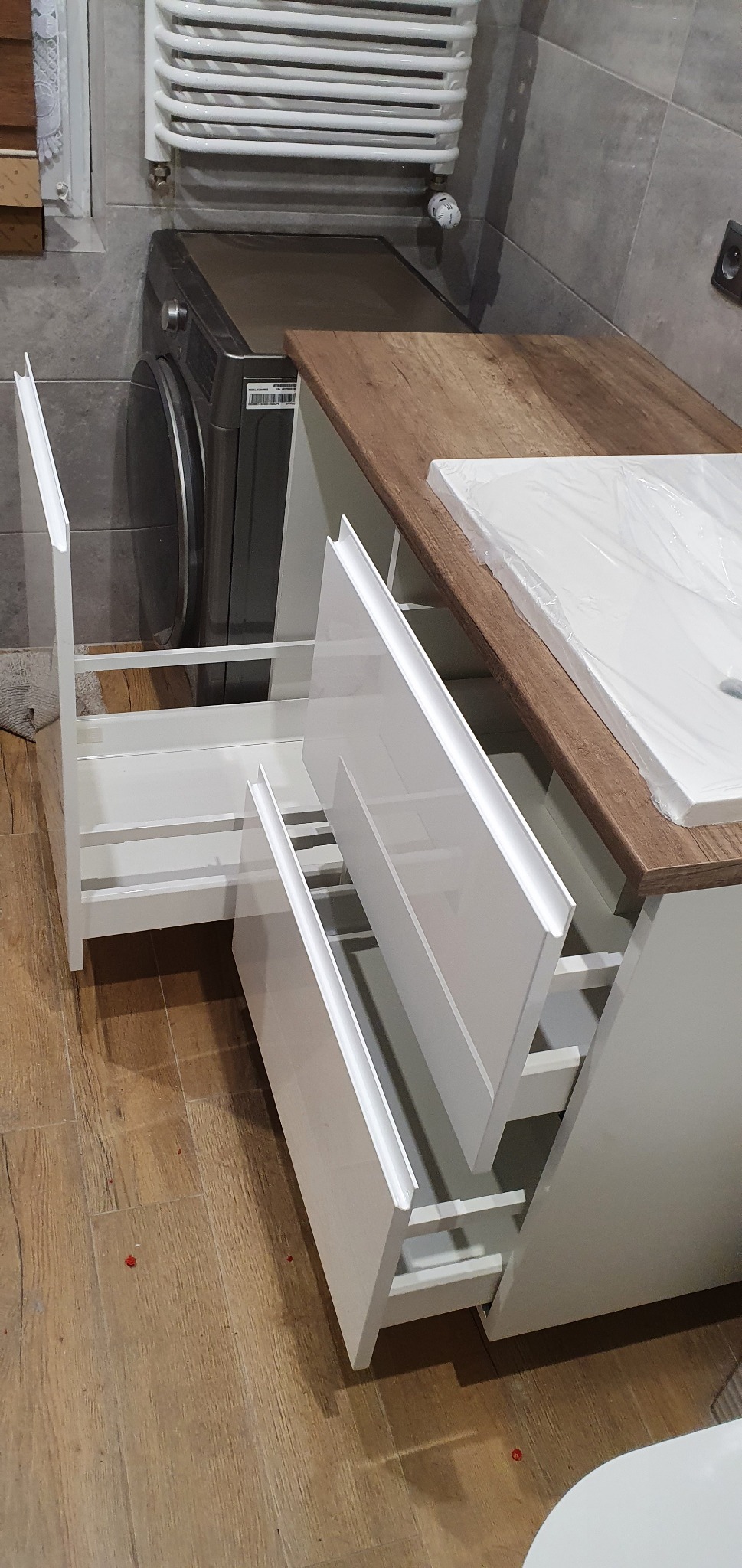 Biała szafka łazienkowa z drewnianym blatem i umywalką w folii, z wysuniętymi szufladami, na tle szarej ściany i pralki.