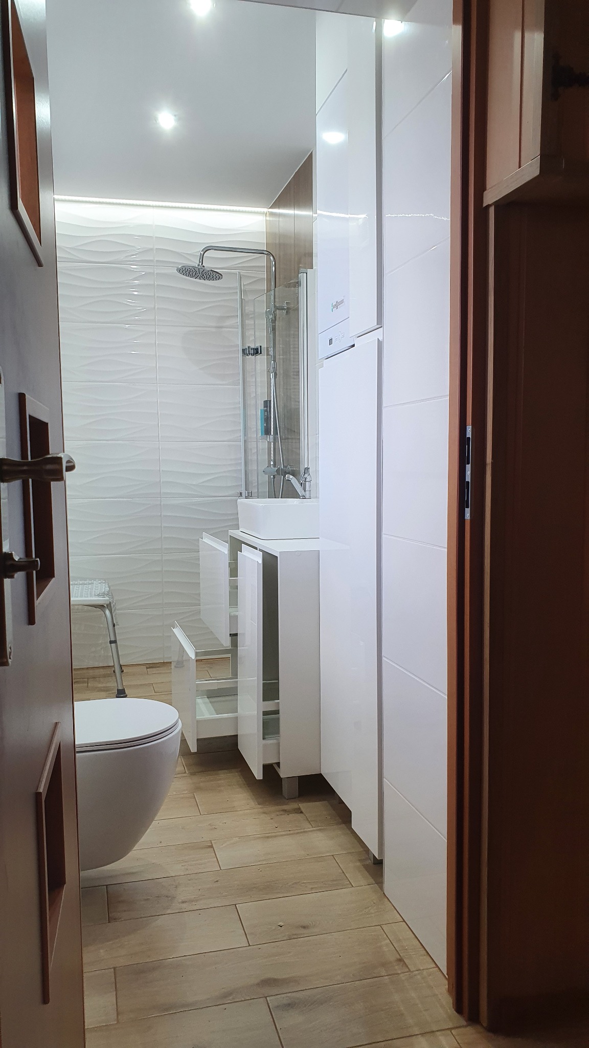 Nowoczesna łazienka z białą zabudową meblową, umywalką nablatową i kabiną prysznicową, widok zza uchylonych drzwi.