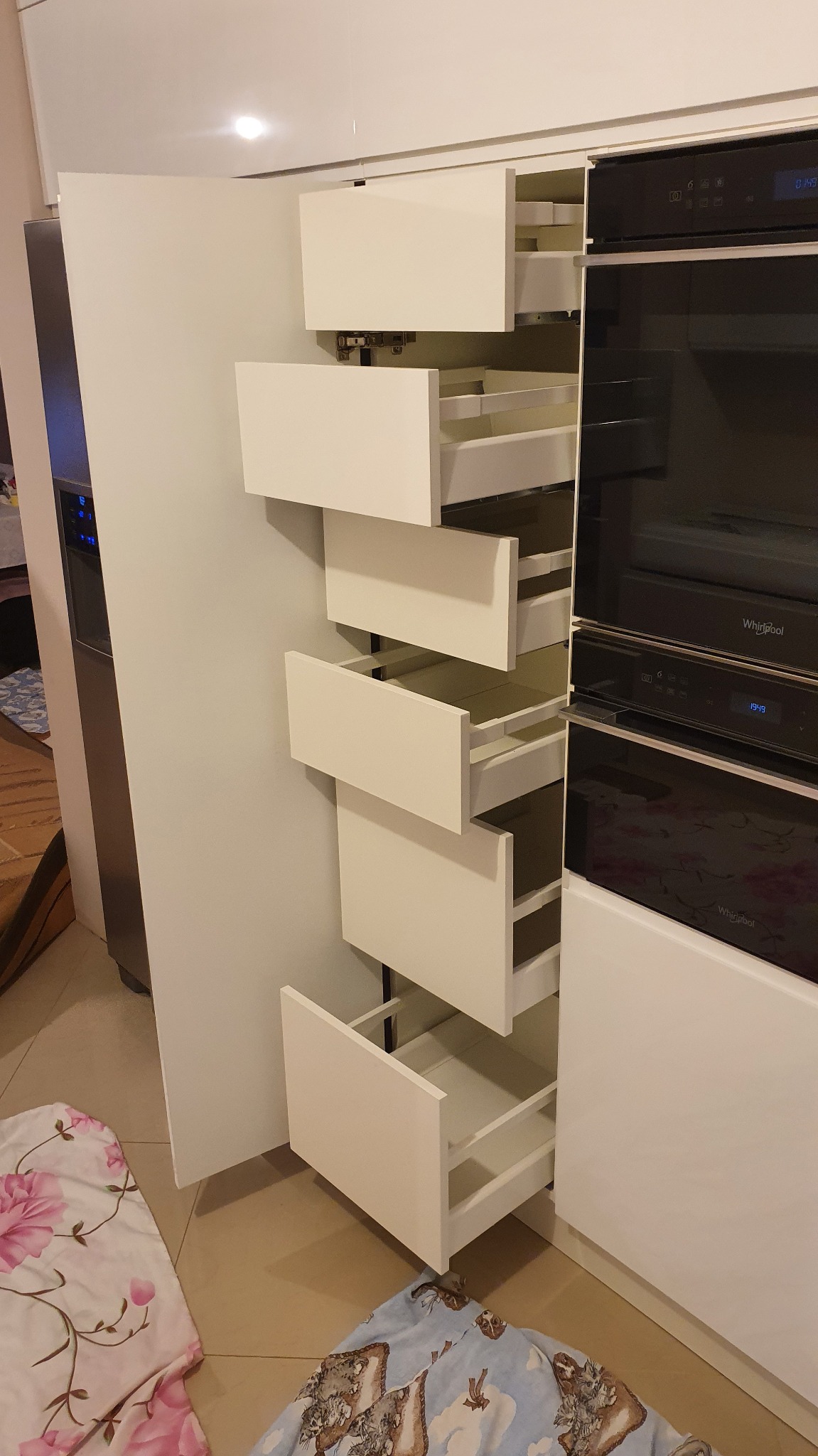 Nowoczesna, biała zabudowa kuchenna z otwartą szafką cargo, ukazującą wysuwane półki. Obok wbudowane piekarniki Whirlpool. Na podłodze fragment wzorzystej tkaniny.