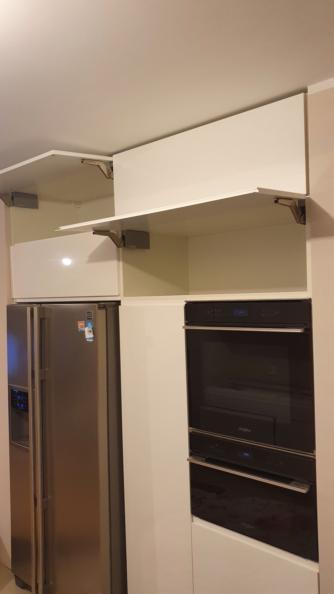 Białe szafki kuchenne z otwartymi frontami, widoczne mechanizmy podnoszące, obok srebrna lodówka i czarny piekarnik w zabudowie.