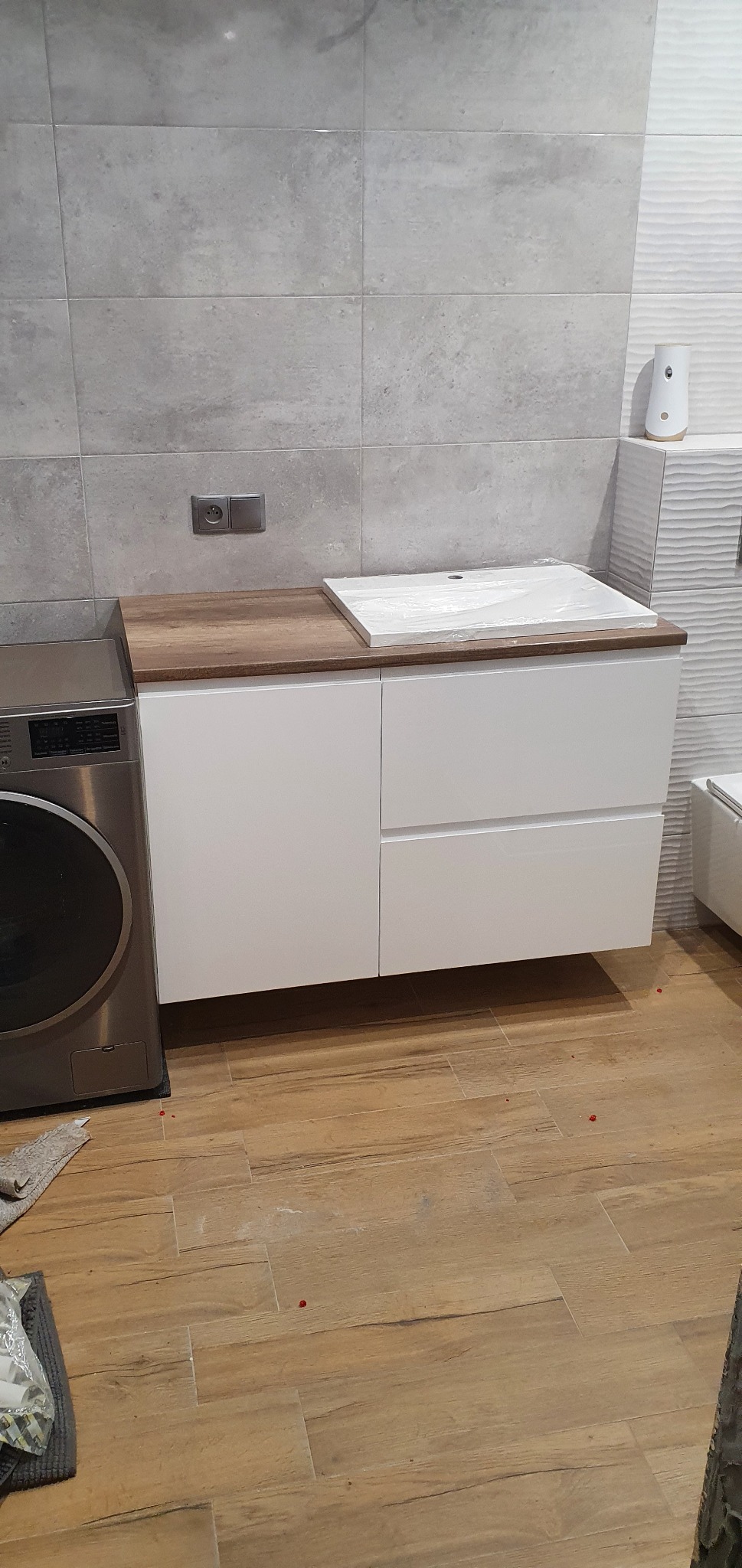 Nowoczesna łazienka z białą szafką pod umywalkę z drewnianym blatem, umywalka jeszcze w folii, obok pralka, szare płytki na ścianie i drewnopodobna podłoga.