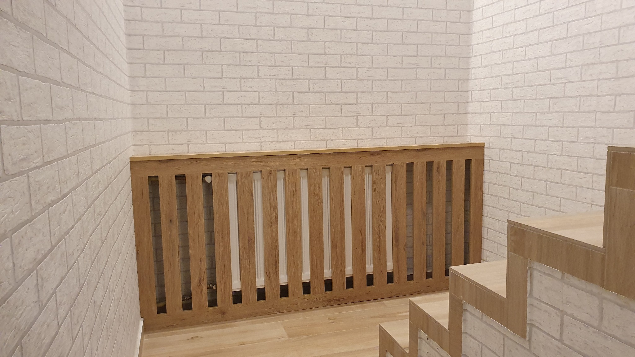 Drewniana zabudowa grzejnika z pionowymi szczebelkami, dopasowana kolorystycznie do drewnianych schodów i podłogi, na tle ściany z białej cegły.