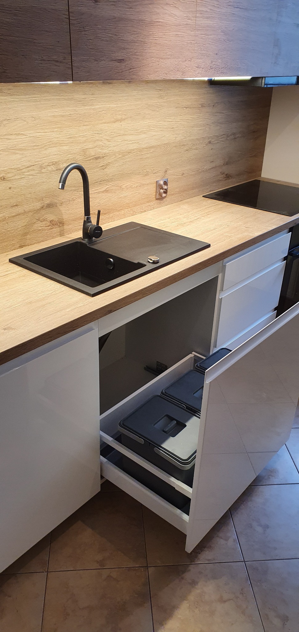 Nowoczesna kuchnia z drewnianym blatem, czarnym zlewem i wysuwanym koszem na śmieci w szafce.