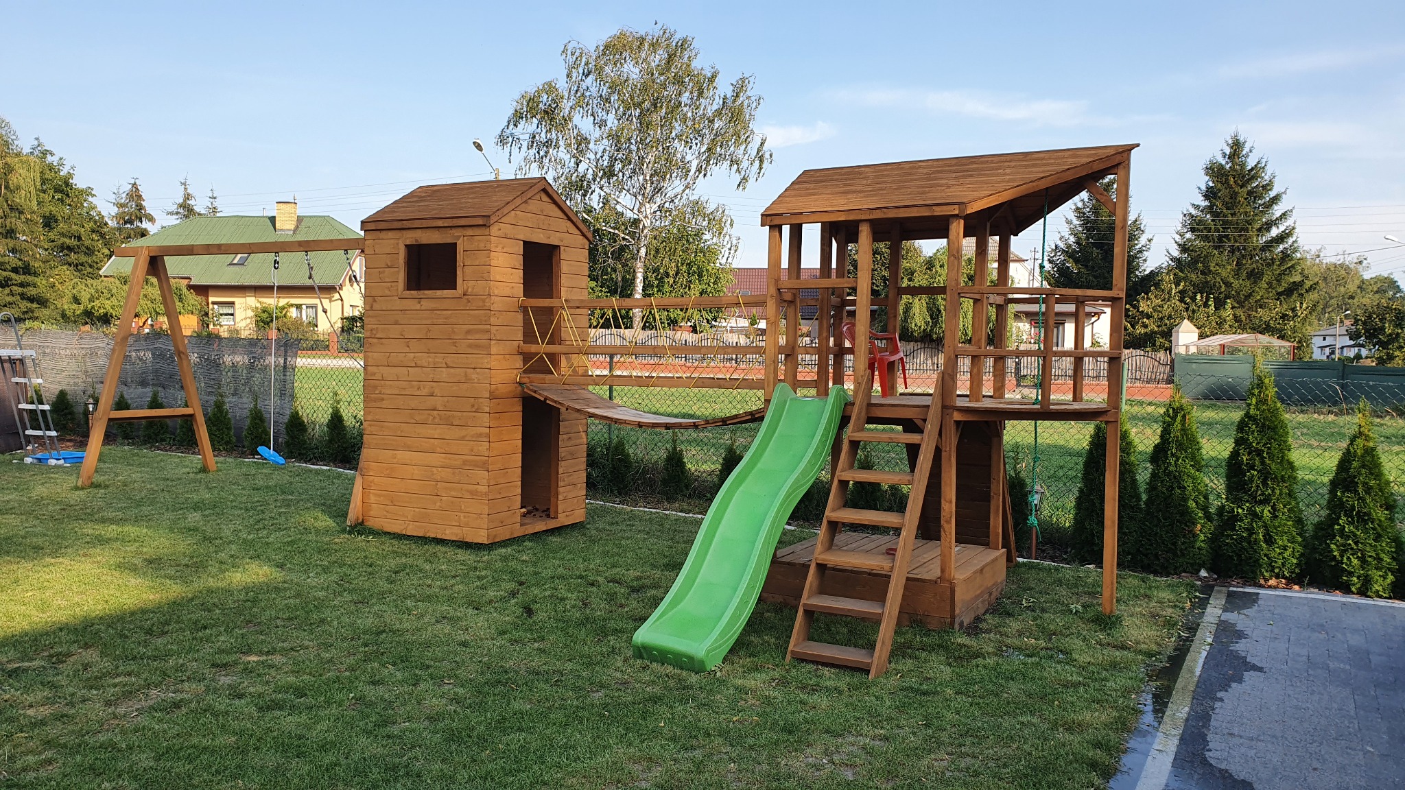 Drewniany plac zabaw z wieżą, zjeżdżalnią i huśtawką na zielonym trawniku, w tle domy i drzewa.