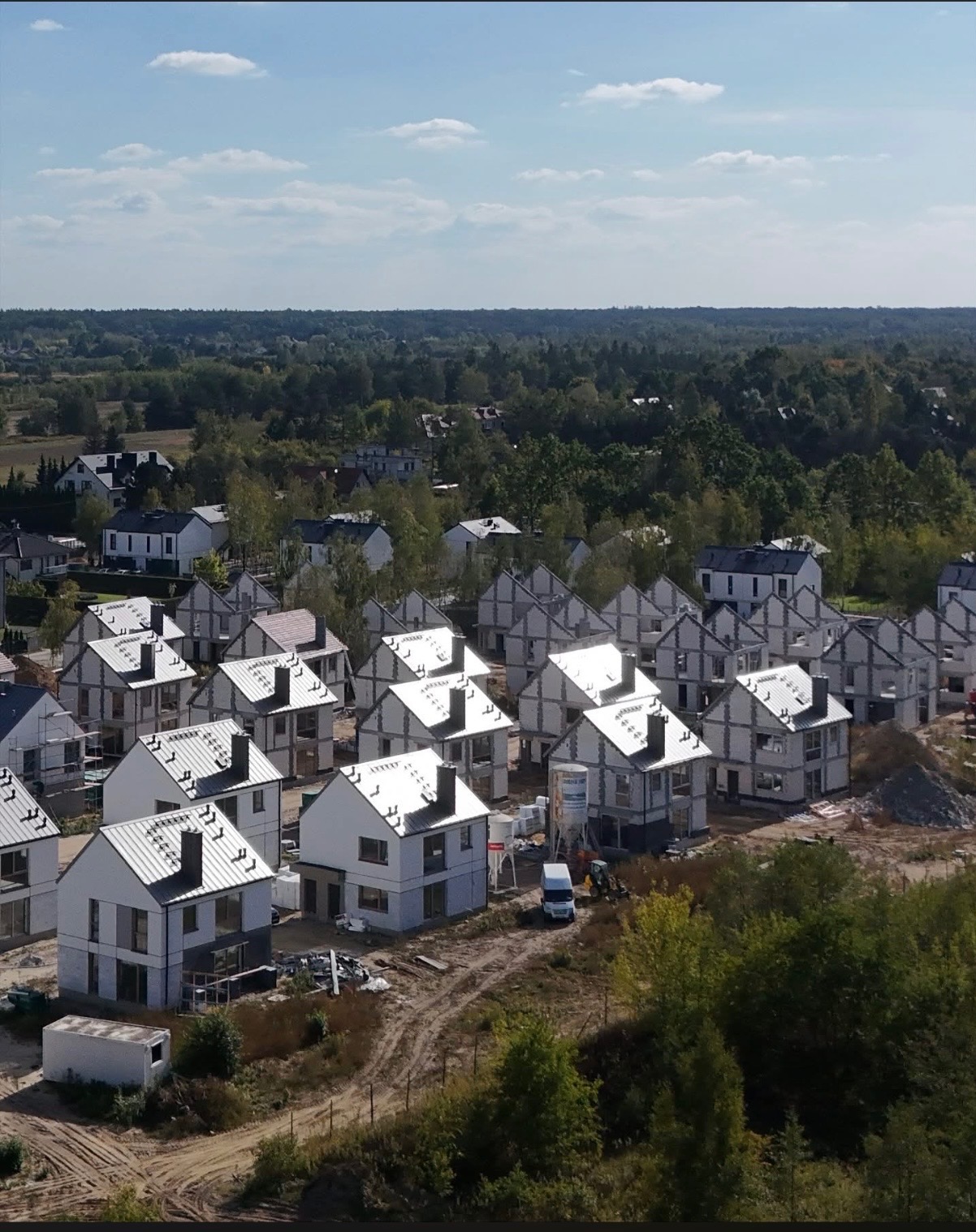 Widok z lotu ptaka na osiedle w budowie z białymi domami w stanie surowym zamkniętym, z widocznymi elementami konstrukcyjnymi i maszynami budowlanymi.