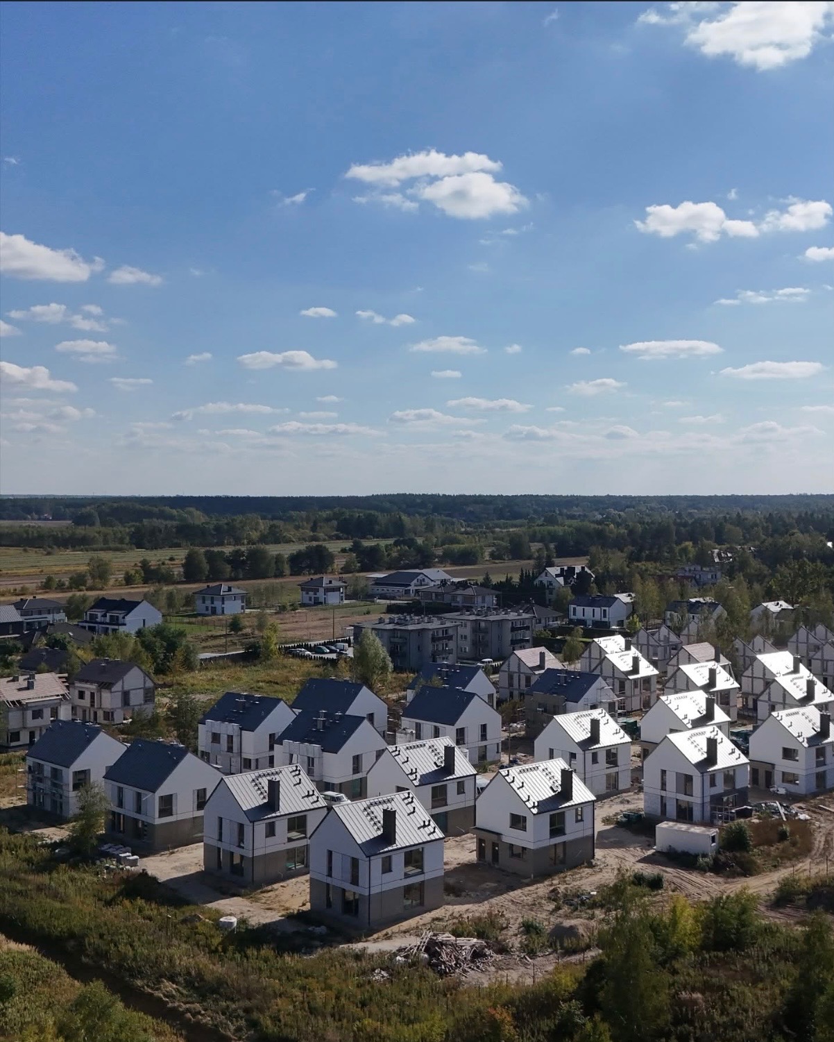 Osiedle domów jednorodzinnych w trakcie budowy, widok z lotu ptaka, błękitne niebo z chmurami w tle. Domy z szarymi dachami i białymi ścianami.