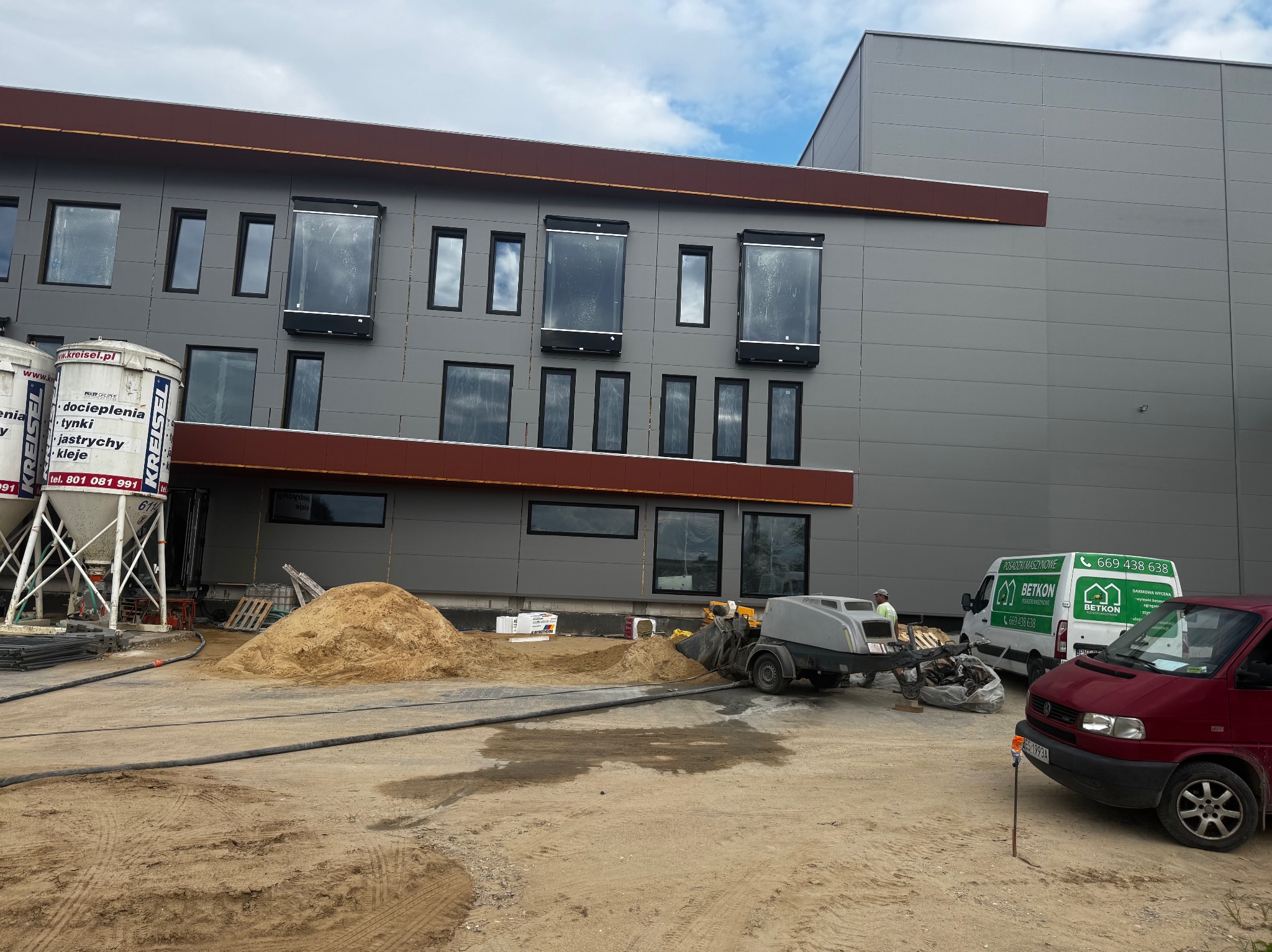 Budynek w trakcie prac z maszyną do wylewania posadzek, stertą piasku i zaparkowanym vanem firmy. Widoczne silosy z materiałami budowlanymi.
