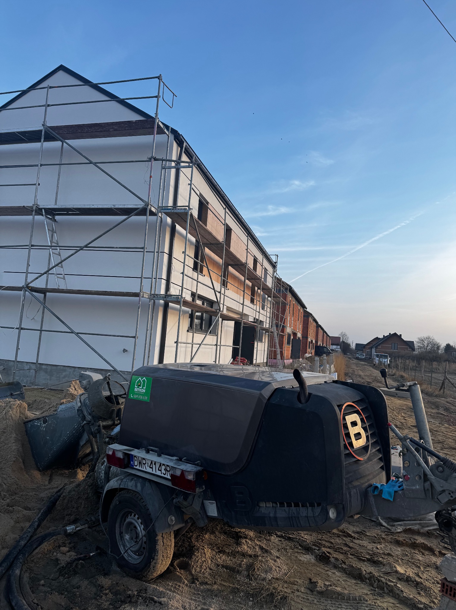 Maszyna do wylewania posadzek na tle budynków w trakcie budowy, z widocznymi rusztowaniami i elementami konstrukcyjnymi. Błękitne niebo w tle.