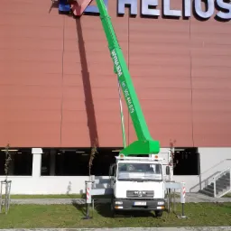 Zielony podnośnik koszowy MAN na trawniku przed budynkiem z napisem HELIOS, dwóch pracowników w koszu montuje logo.