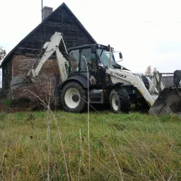 Traktor z łyżką marki Terex kopie ziemię obok starego, ceglanego budynku na zarośniętym polu.