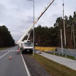 Biały dźwig samochodowy PAC podnosi betonowy słup na poboczu drogi w pobliżu lasu, zabezpieczony barierkami i pachołkami.