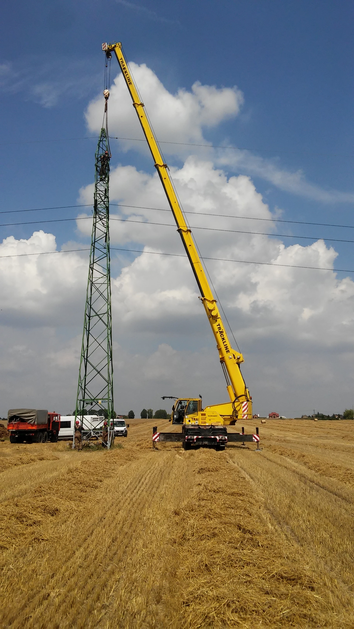 Żółty dźwig teleskopowy Trackline podnosi pracowników na szczyt metalowej konstrukcji słupa energetycznego na tle błękitnego nieba z chmurami, na polu po żniwach.