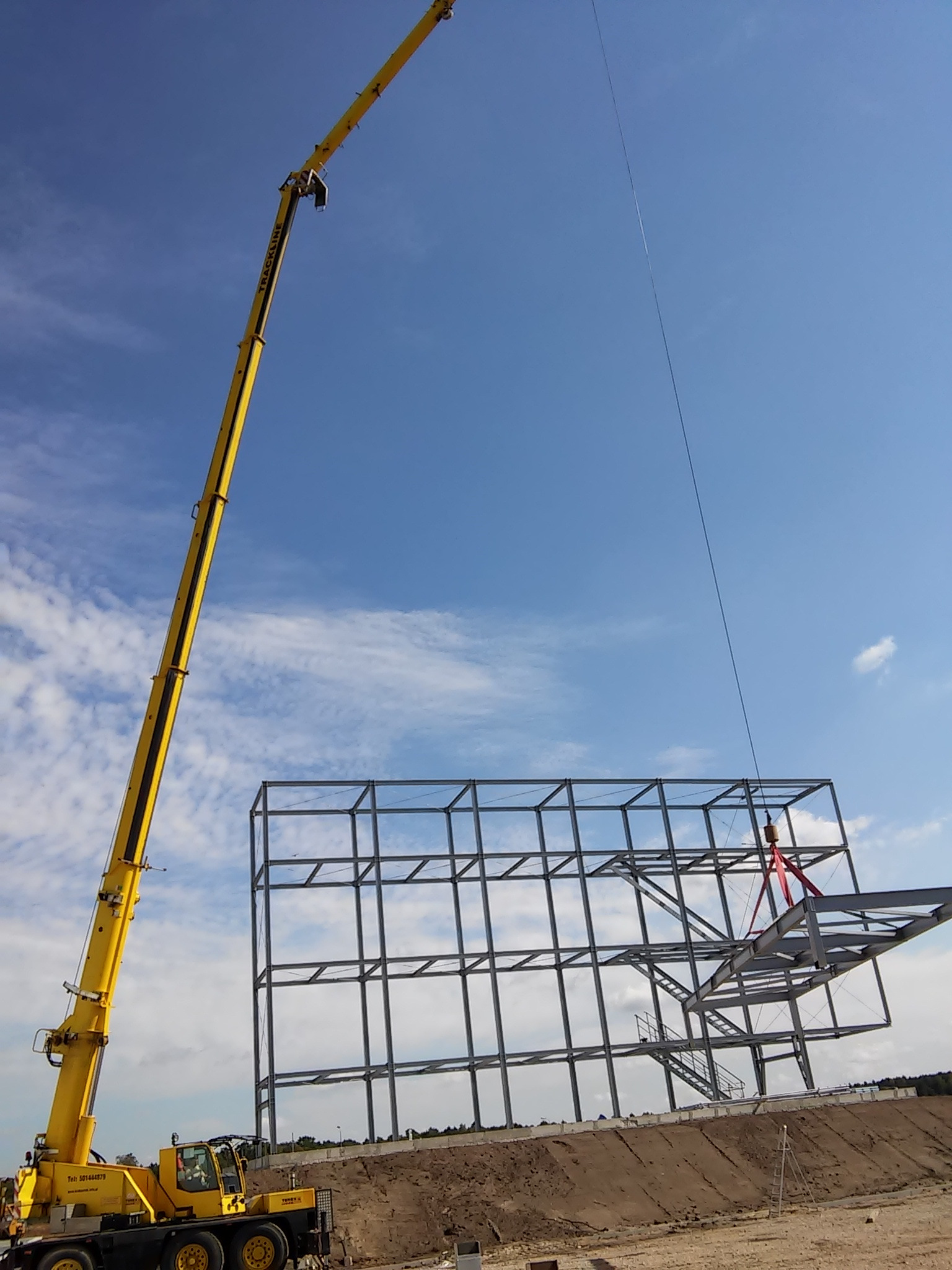 Żółty dźwig teleskopowy podnosi stalową konstrukcję budynku na placu budowy pod błękitnym niebem z chmurami.