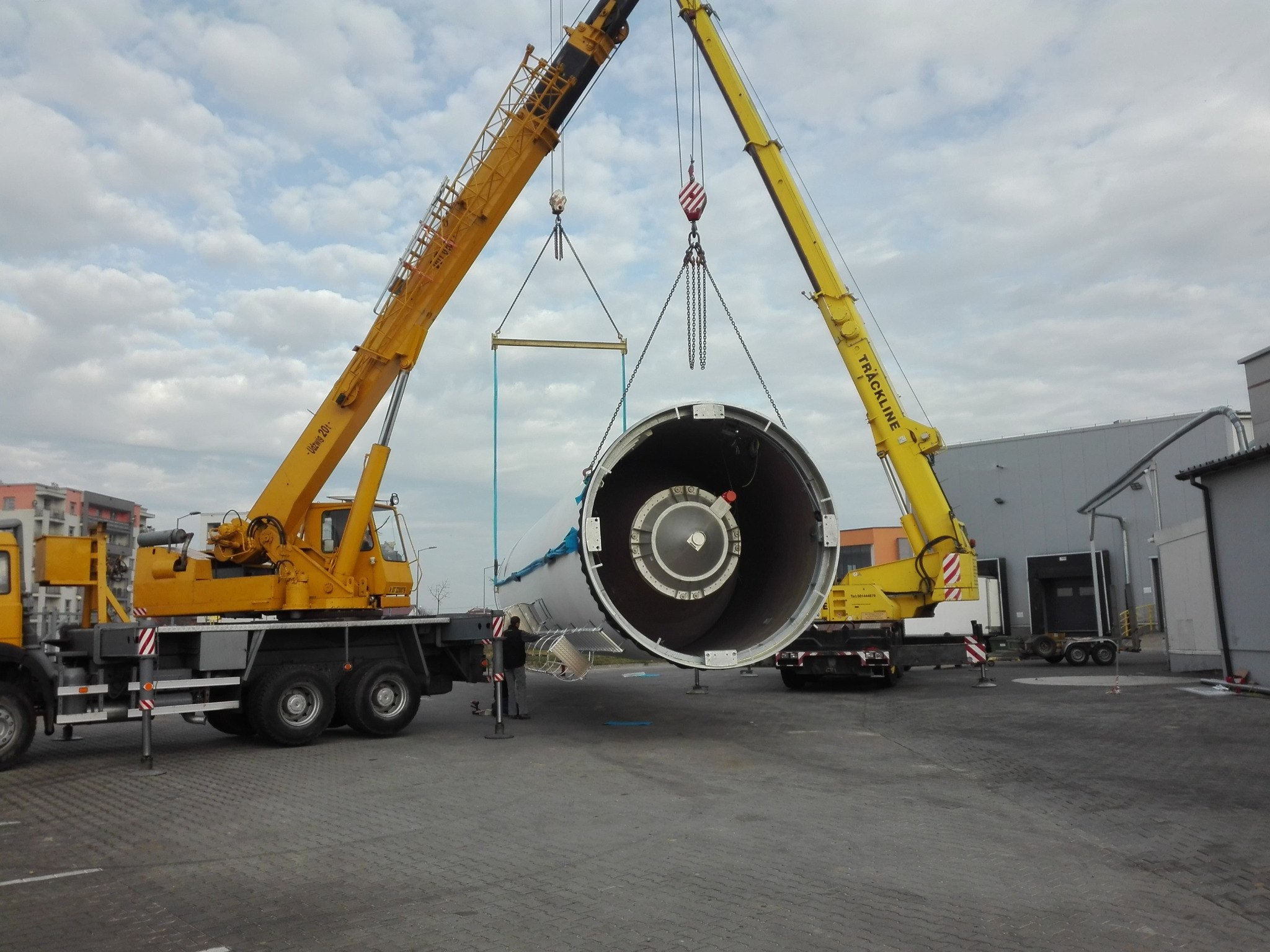 Dwa żółte dźwigi samochodowe podnoszą element turbiny wiatrowej na placu budowy, widoczna osoba nadzorująca operację.
