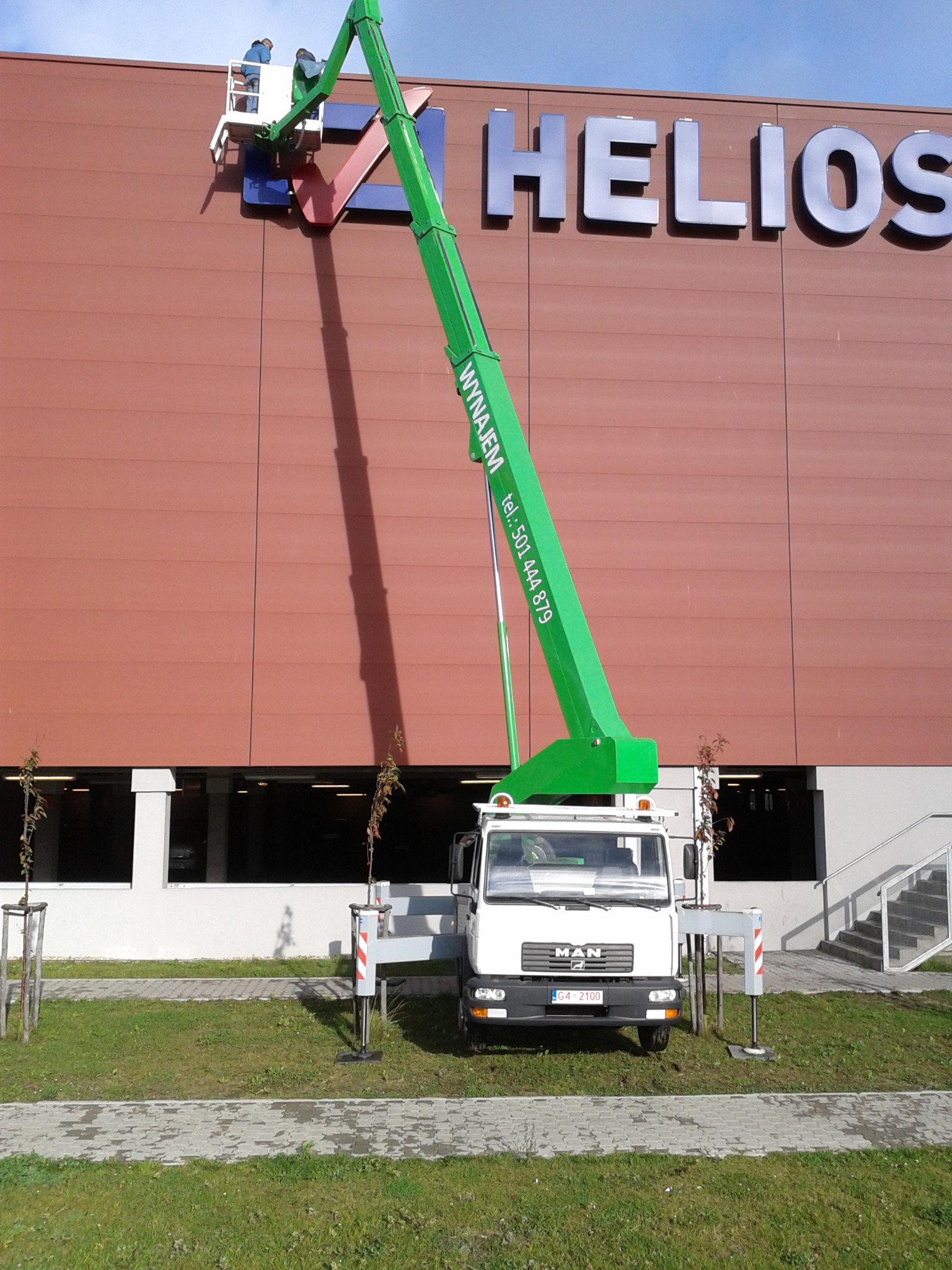 Zielony podnośnik koszowy MAN na trawniku przed budynkiem z napisem HELIOS, dwóch pracowników w koszu montuje logo.