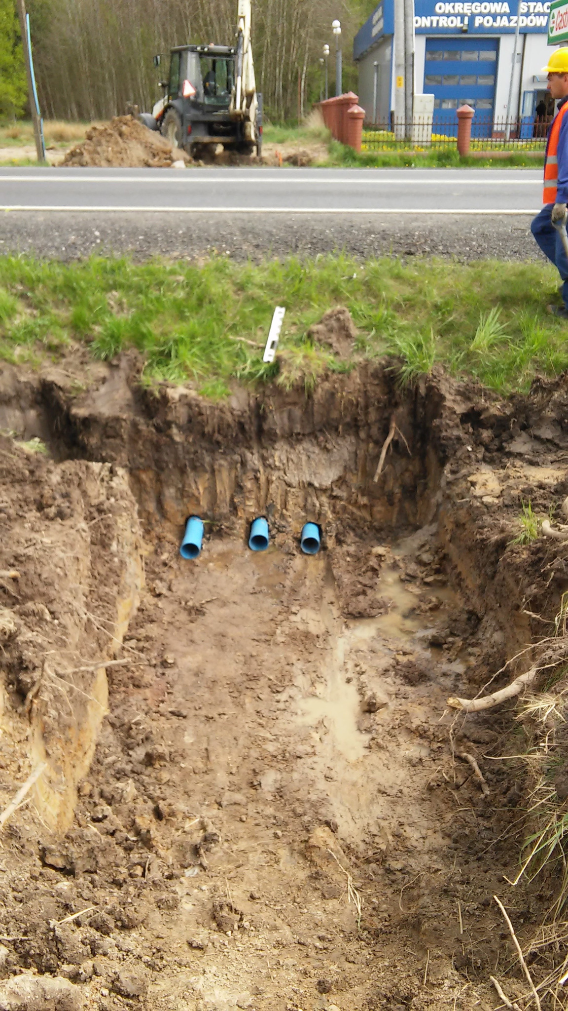 Wykop w ziemi z trzema niebieskimi rurami ułożonymi poziomo, w tle koparka i pracownik w żółtym kasku przy drodze.