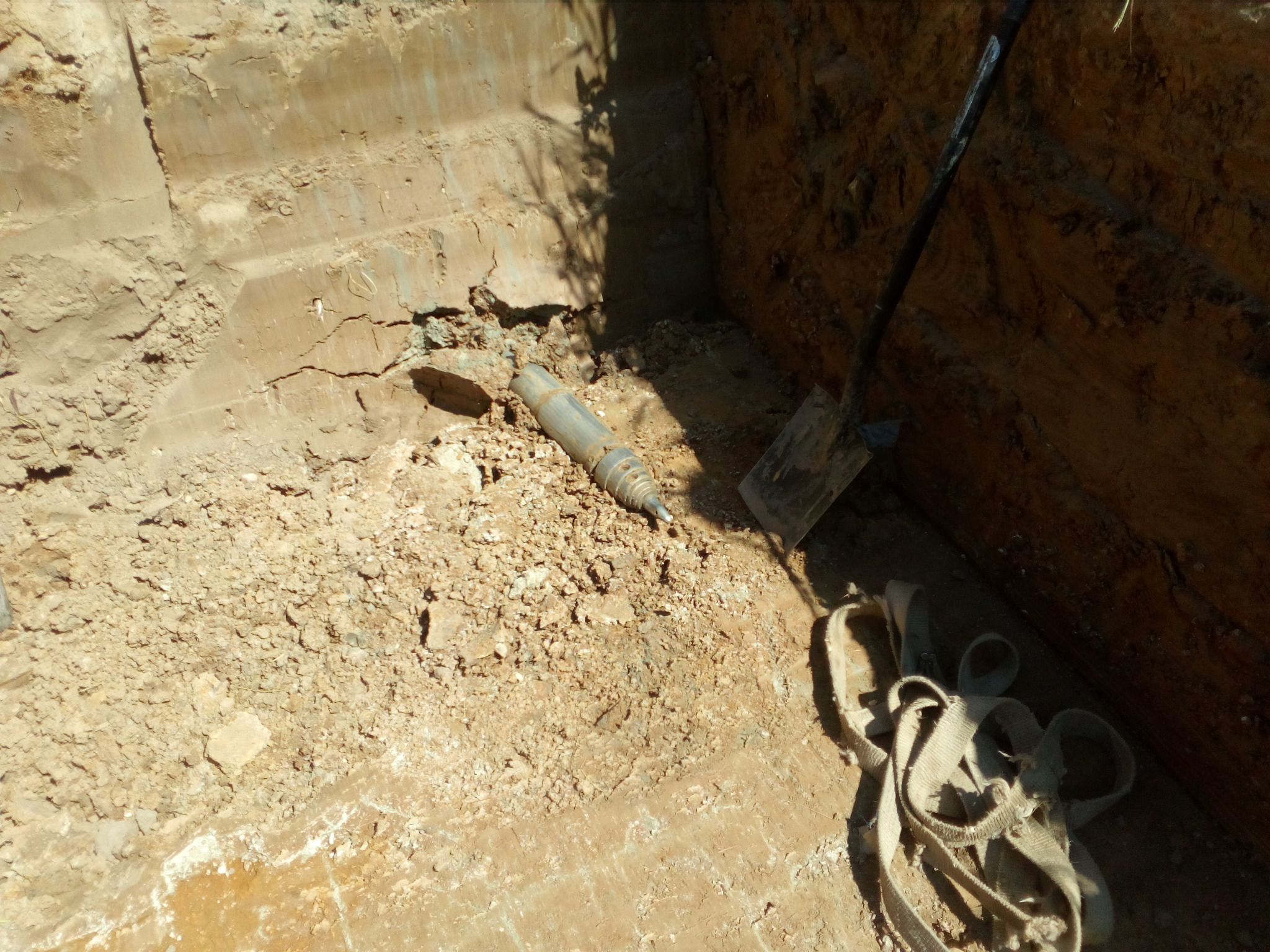 Wykop w ziemi z widoczną rurą, łopatą i pasami transportowymi na dnie wykopu.