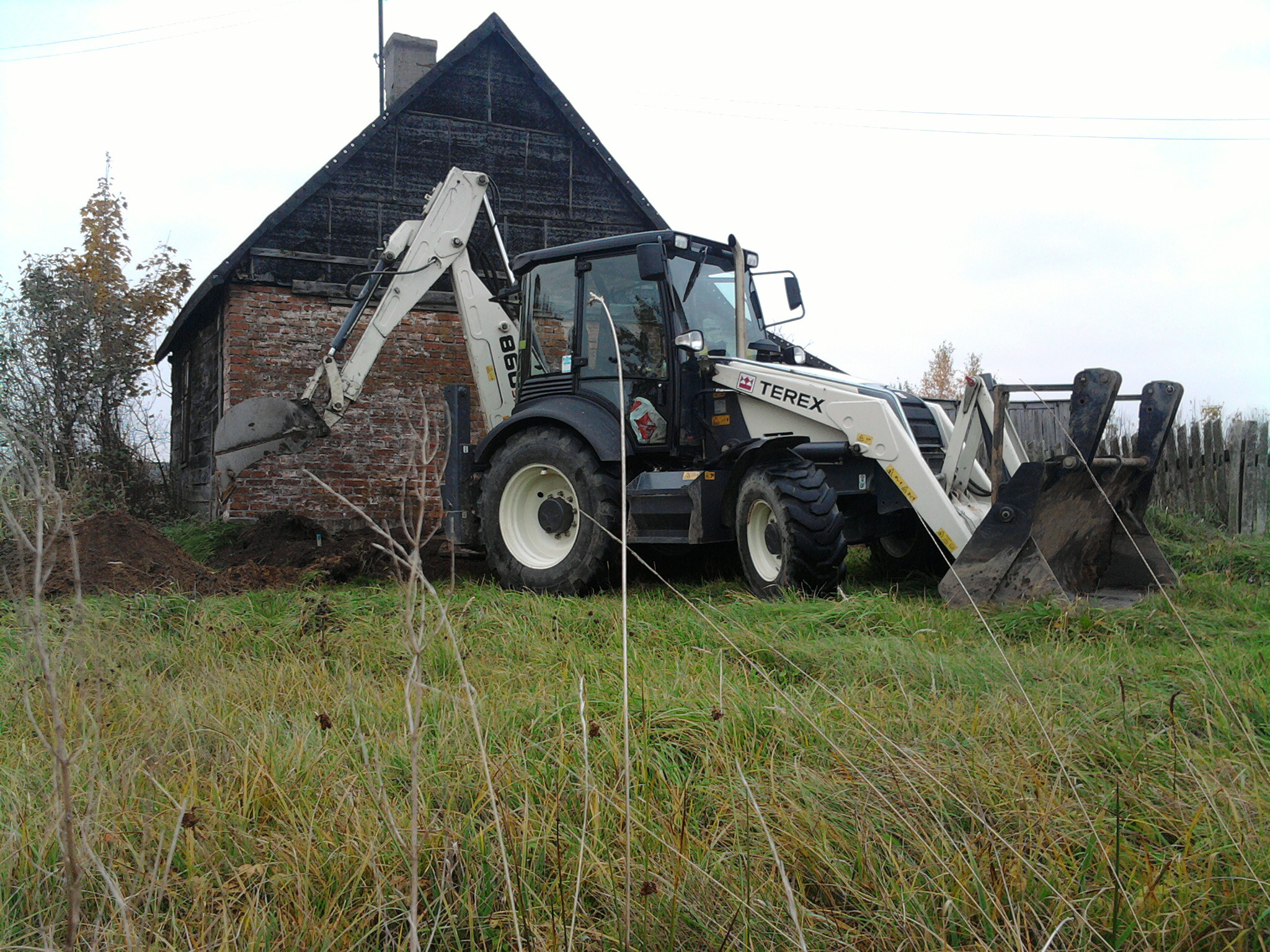 Traktor z łyżką marki Terex kopie ziemię obok starego, ceglanego budynku na zarośniętym polu.
