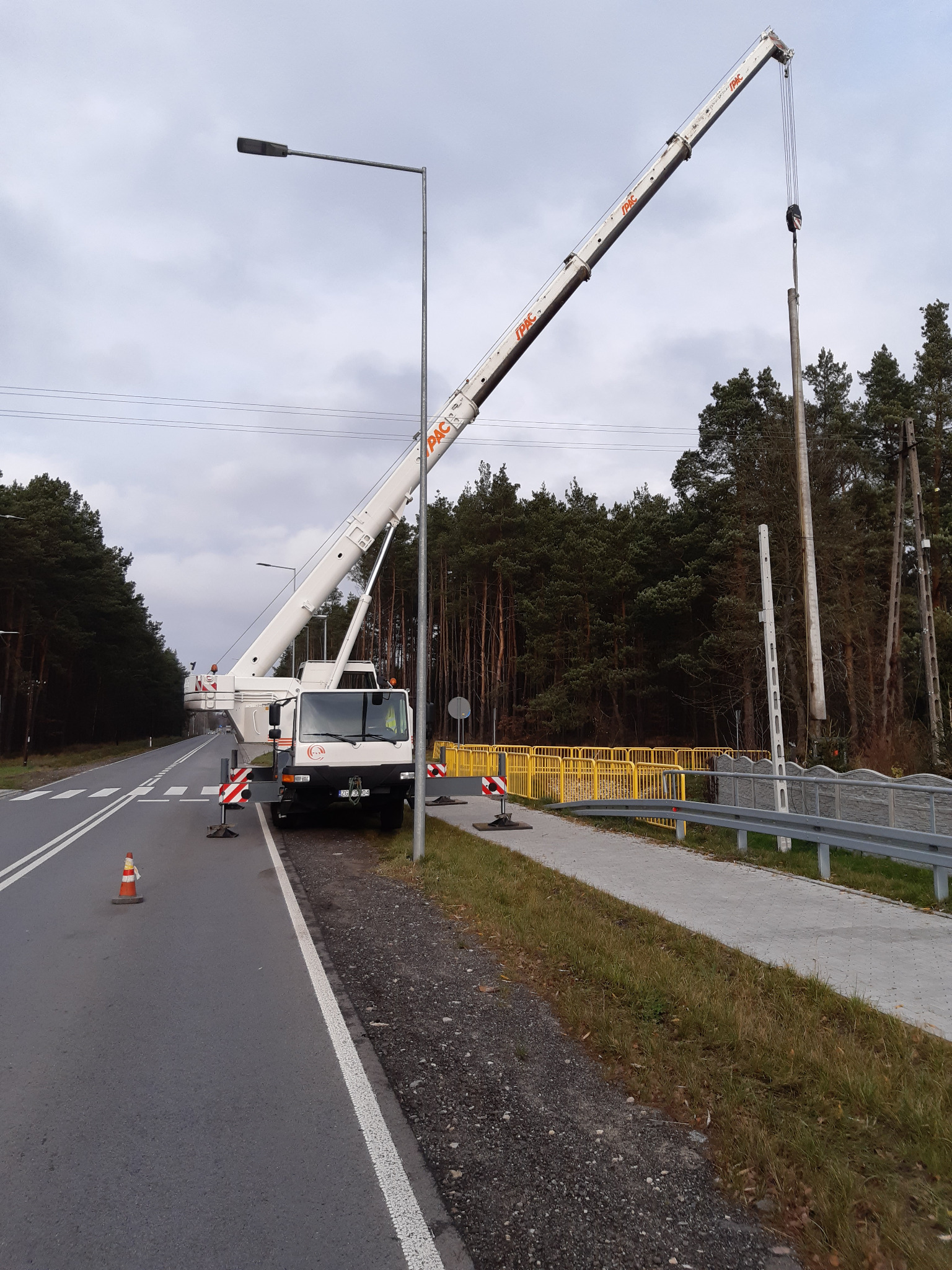 Biały dźwig samochodowy PAC podnosi betonowy słup na poboczu drogi w pobliżu lasu, zabezpieczony barierkami i pachołkami.